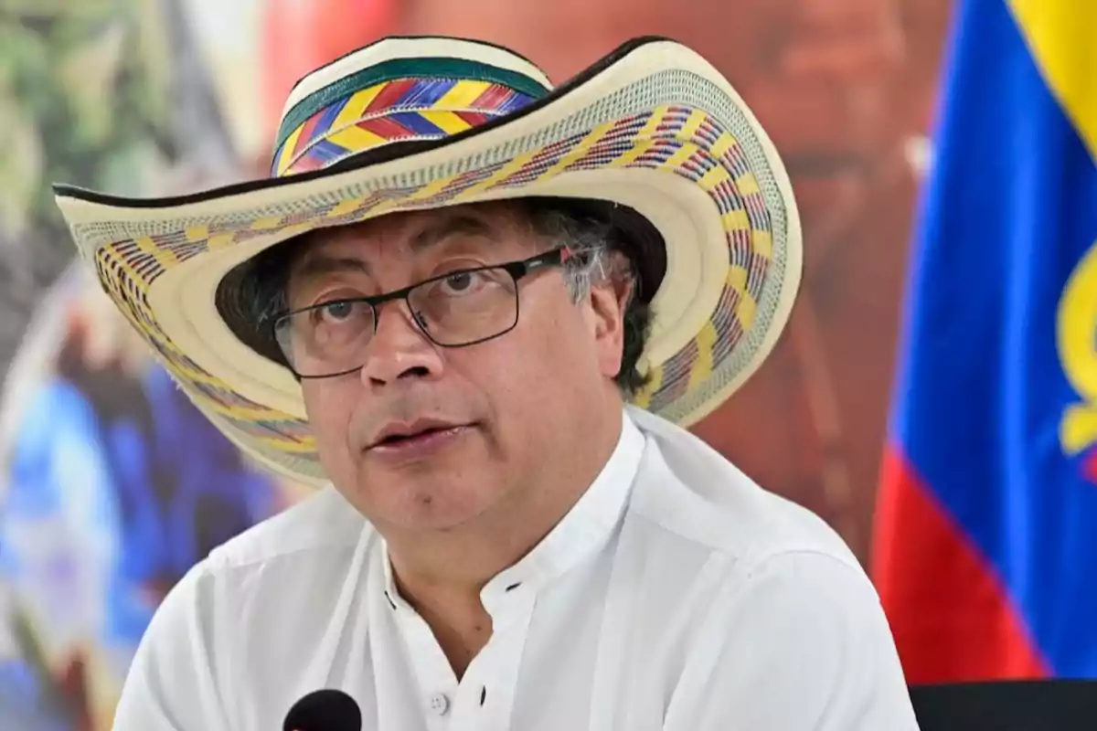 Un hombre con sombrero tradicional y gafas, vistiendo una camisa blanca, frente a un micrófono y una bandera de fondo.