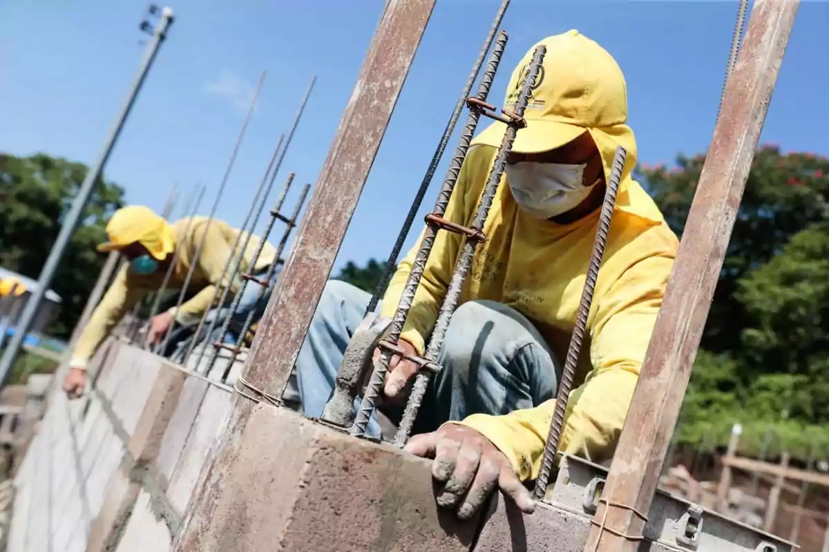 Dos trabajadores de la construcción con ropa amarilla y mascarilla levantan una pared de bloques bajo el sol