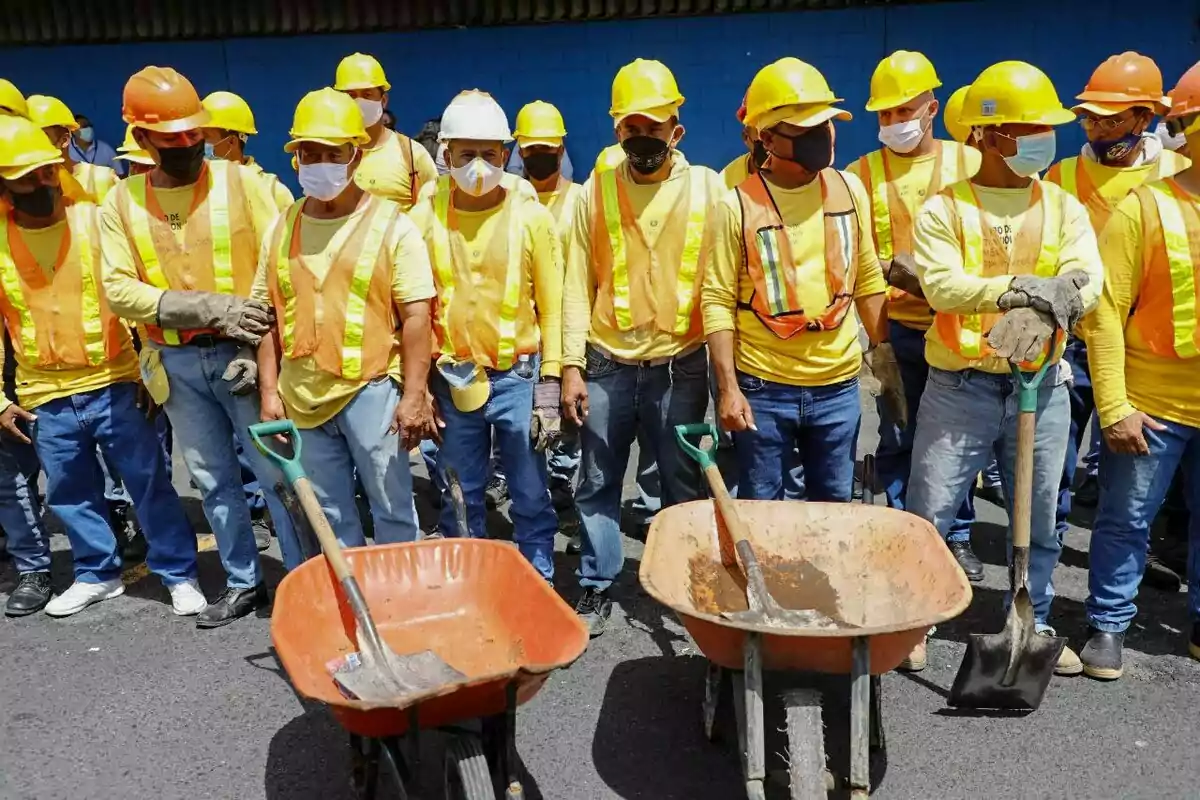 Grupo de trabajadores de la construcción con cascos y chalecos reflectantes de pie junto a carretillas y palas en una calle Grupo de trabajadores de la construcción con cascos y chalecos reflectantes de pie junto a carretillas y palas en una calle