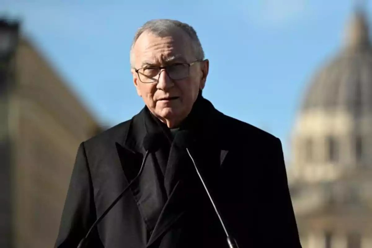 A man with glasses and a dark coat speaks in front of a microphone with a domed building in the background. A man with glasses and a dark coat speaks in front of a microphone with a domed building in the background.