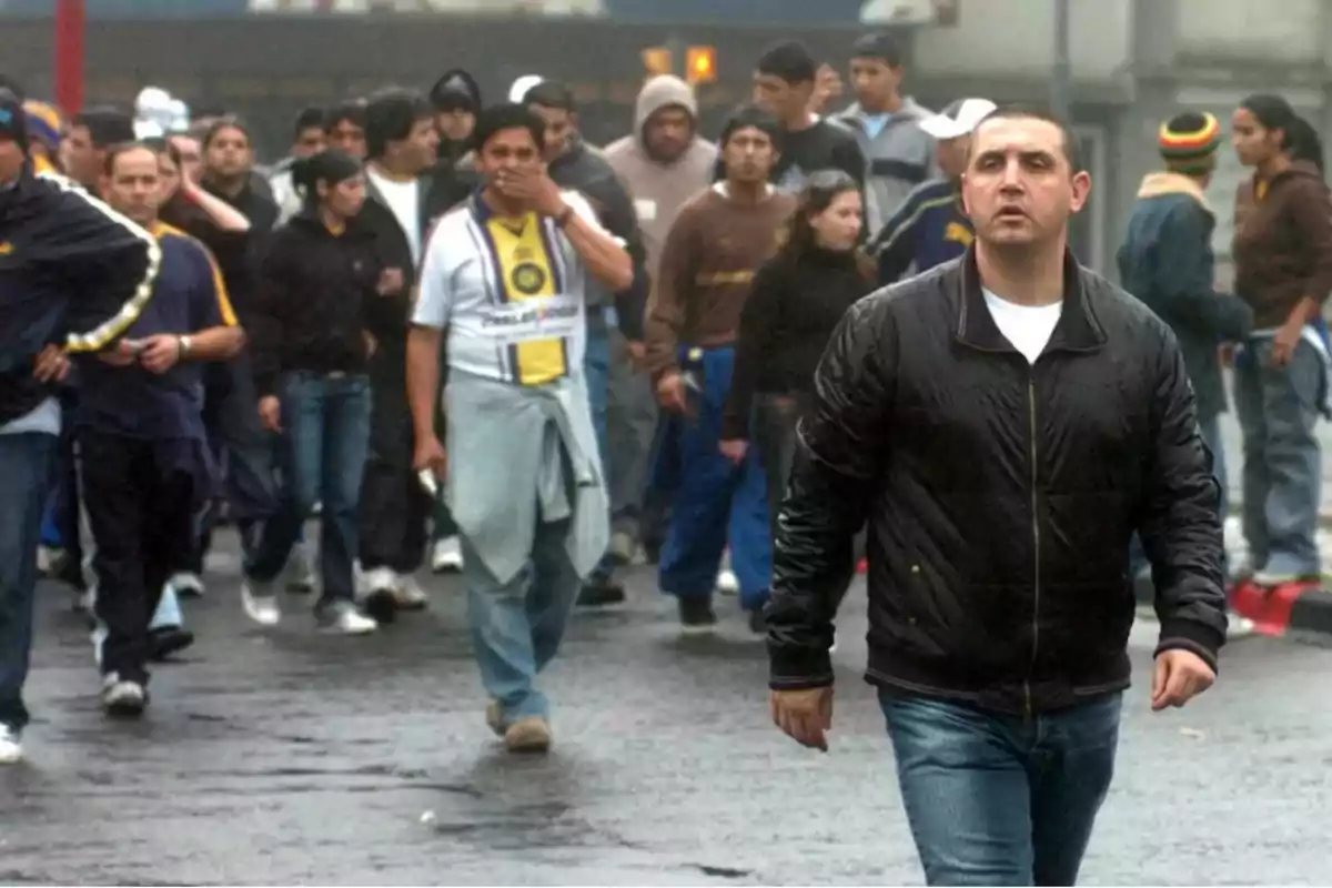 Un grupo de personas camina por una calle mojada, destacando un hombre con chaqueta negra al frente.