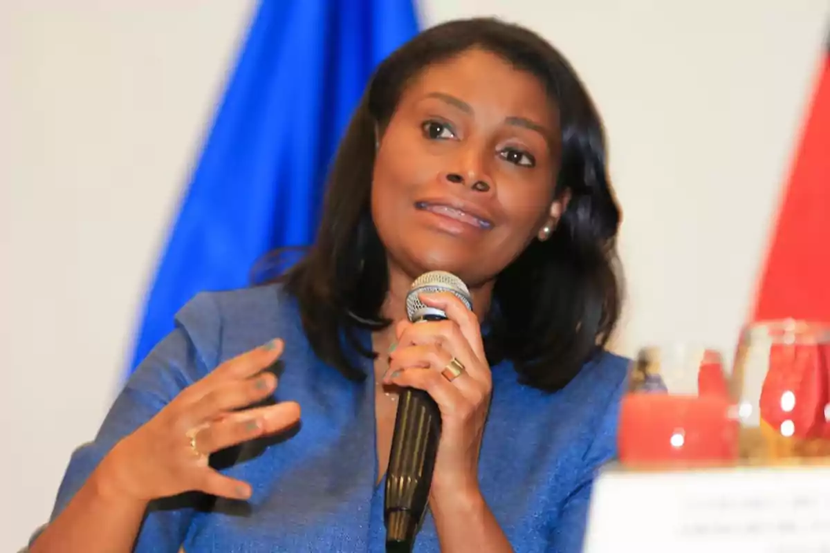 A woman speaking into a microphone with a blue flag in the background.