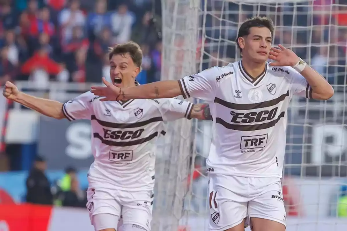 Two players celebrate a goal enthusiastically in a stadium, both wearing white uniforms with brown details, and one of them puts his hand to his ear while the other shouts excitedly. Two players celebrate a goal enthusiastically in a stadium, both wearing white uniforms with brown details, and one of them puts his hand to his ear while the other shouts excitedly.