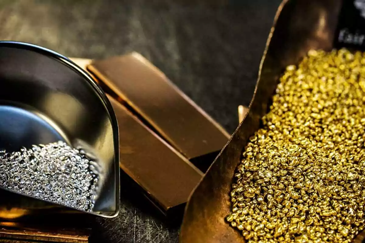 Una bandeja con pequeñas piezas de plata y otra con piezas de oro junto a lingotes de metal sobre una superficie oscura. Una bandeja con pequeñas piezas de plata y otra con piezas de oro junto a lingotes de metal sobre una superficie oscura.