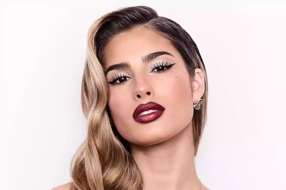 Una persona con maquillaje elegante y labios oscuros posando frente a un fondo blanco.