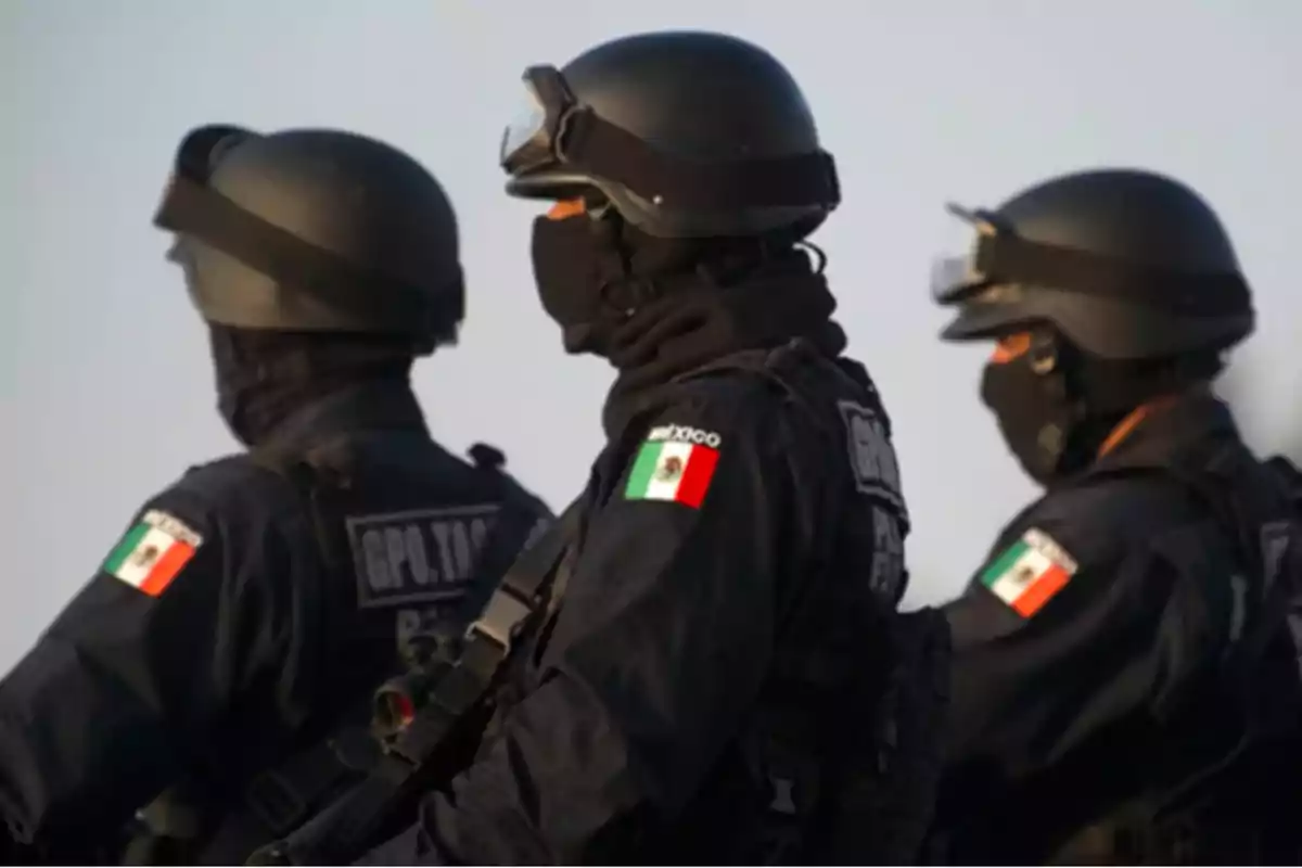 Tres agentes de policía con uniformes oscuros y cascos, portando la bandera de México en sus brazos, observan en la misma dirección.