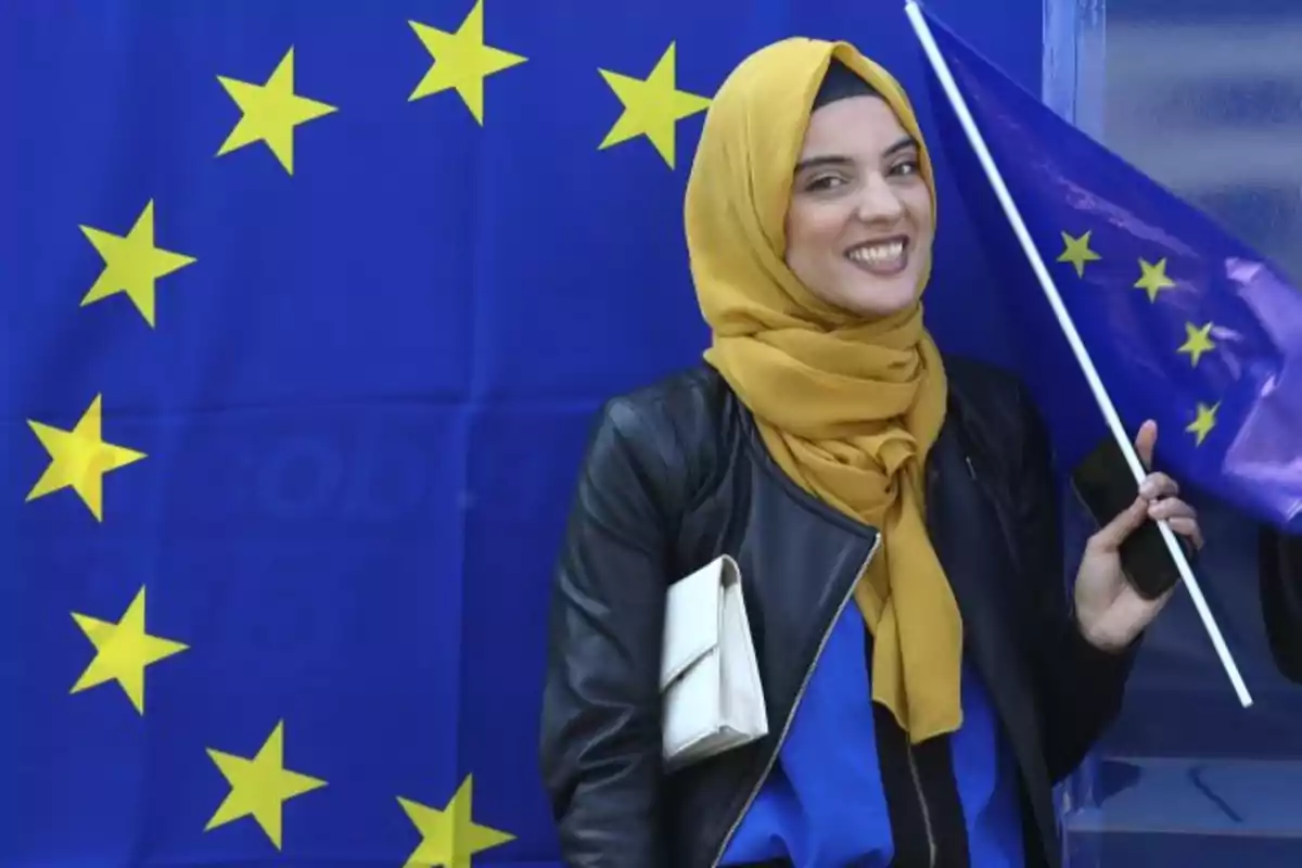 Woman wearing a yellow hijab smiles while holding a European Union flag in front of a blue background with yellow stars. Woman wearing a yellow hijab smiles while holding a European Union flag in front of a blue background with yellow stars.