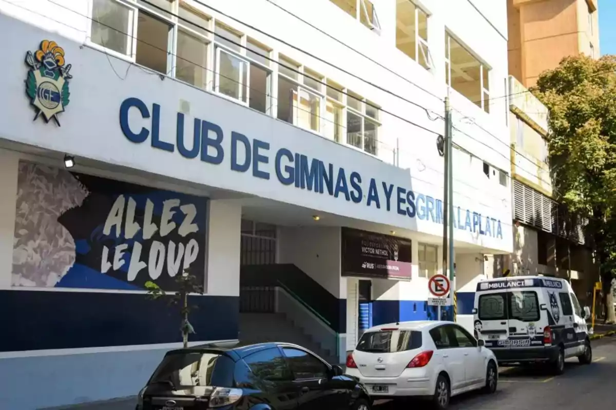Facade of a building with the sign "Club de Gimnasia y Esgrima La Plata" and several vehicles parked on the street.