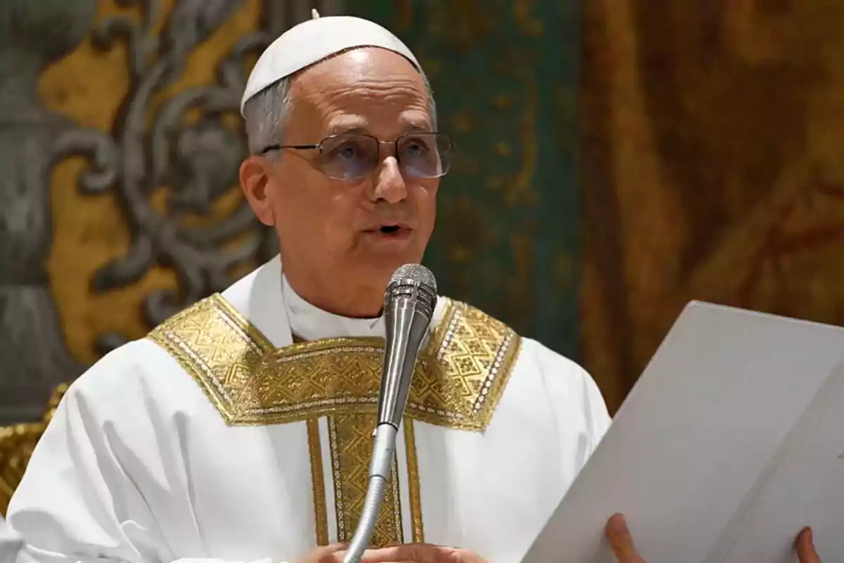 Un hombre con vestimenta religiosa blanca y dorada sostiene un micrófono y un documento mientras habla.