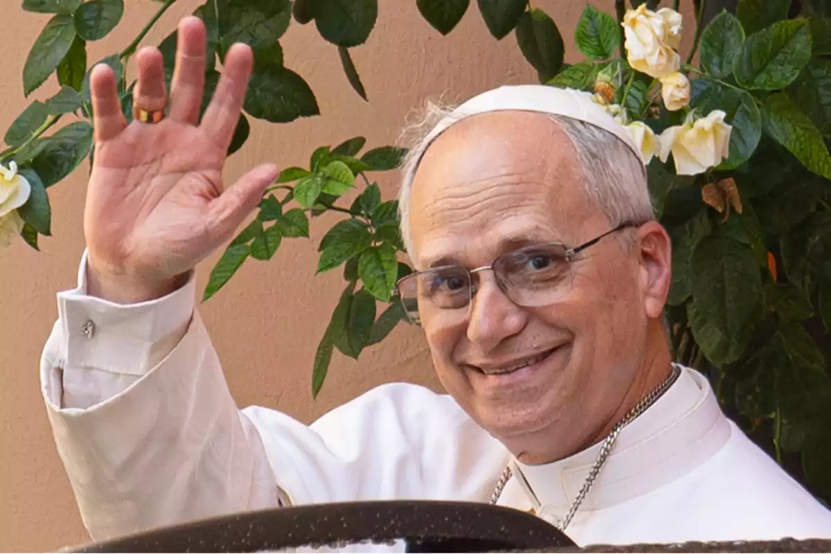 Un hombre con vestimenta religiosa blanca y solideo sonríe y saluda con la mano levantada frente a un fondo de plantas con flores blancas. Un hombre con vestimenta religiosa blanca y solideo sonríe y saluda con la mano levantada frente a un fondo de plantas con flores blancas.
