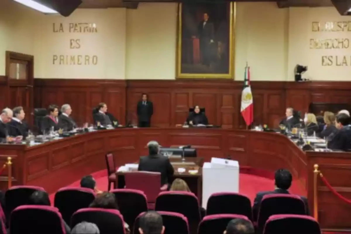 Una sala de tribunal con jueces sentados en un estrado semicircular, un retrato en la pared y una bandera nacional.