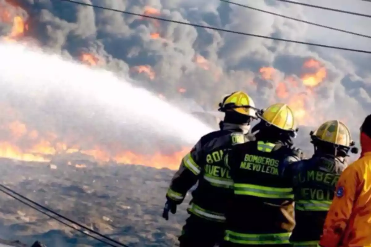 Bomberos combatiendo un incendio con mangueras mientras el fuego y el humo se elevan al fondo.