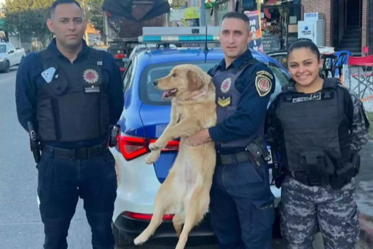 Three police officers pose smiling next to a beige dog in front of a patrol car on the street.