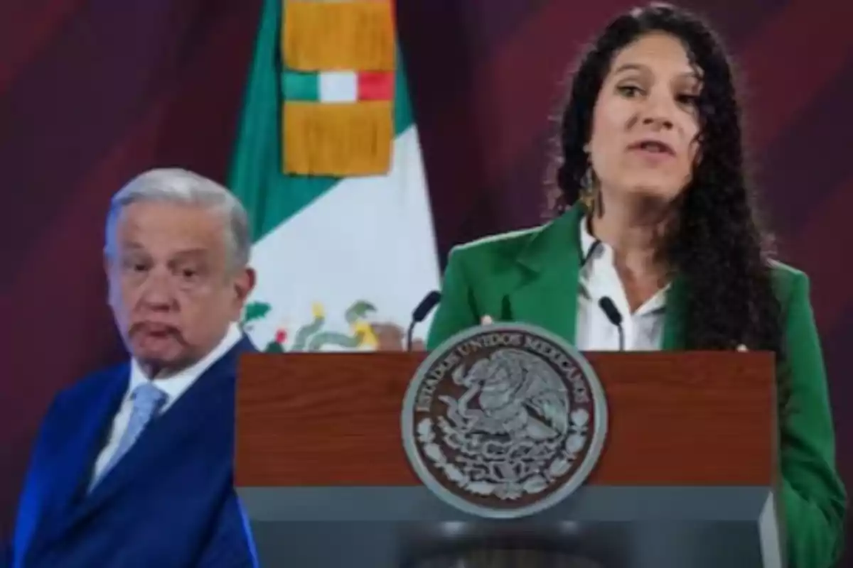 Una mujer hablando en un podio con el escudo de México mientras un hombre mayor está de pie a su lado, con una bandera mexicana de fondo.