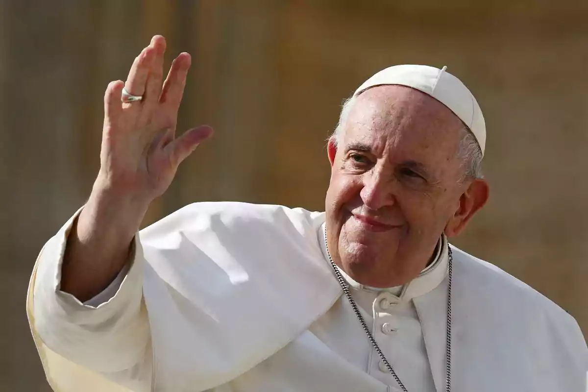 An elderly man in white religious attire and a small hat waves with a raised hand and a friendly expression. An elderly man in white religious attire and a small hat waves with a raised hand and a friendly expression.
