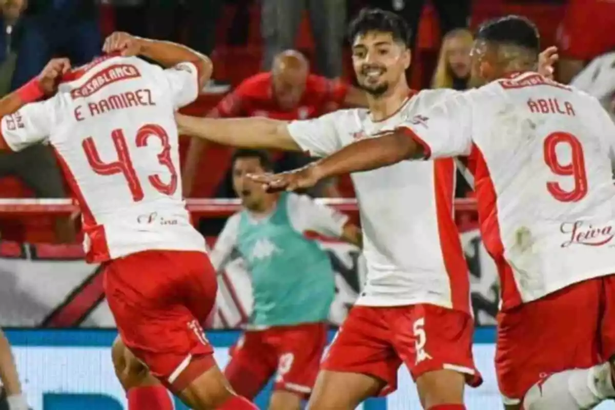 Three players in white and red uniforms celebrate a goal on the field while other players and fans are visible in the background. Three players in white and red uniforms celebrate a goal on the field while other players and fans are visible in the background.