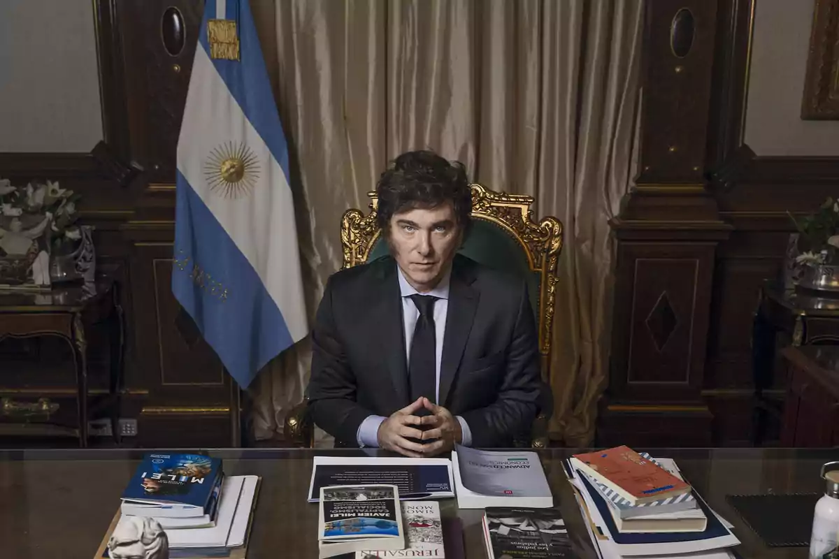 Un hombre sentado en un escritorio con libros y documentos, frente a una bandera de Argentina.