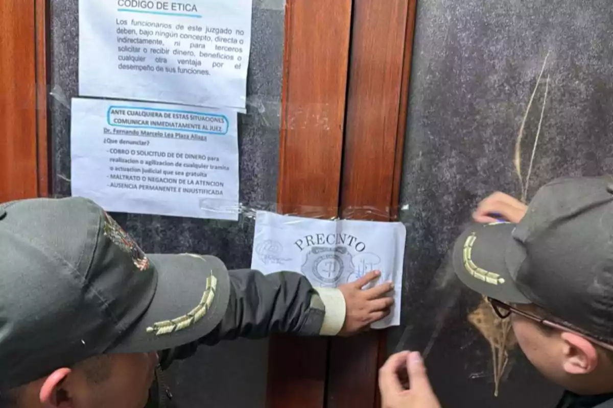 Dos personas con uniforme policial colocan un sello de precinto en la puerta de madera de un edificio junto a avisos informativos pegados en la pared. Dos personas con uniforme policial colocan un sello de precinto en la puerta de madera de un edificio junto a avisos informativos pegados en la pared.