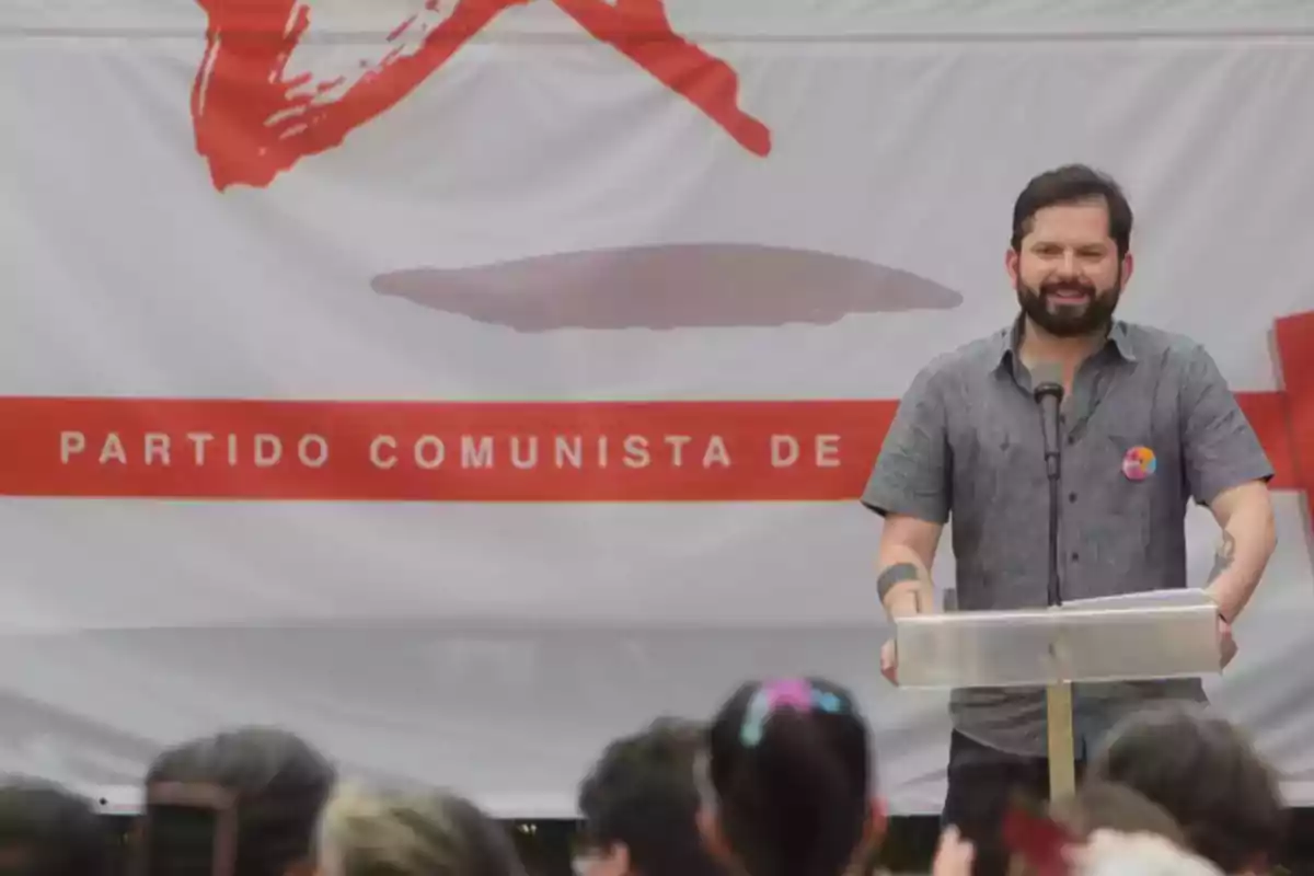 Un hombre de pie frente a un podio con un micrófono, con un cartel del Partido Comunista de fondo.