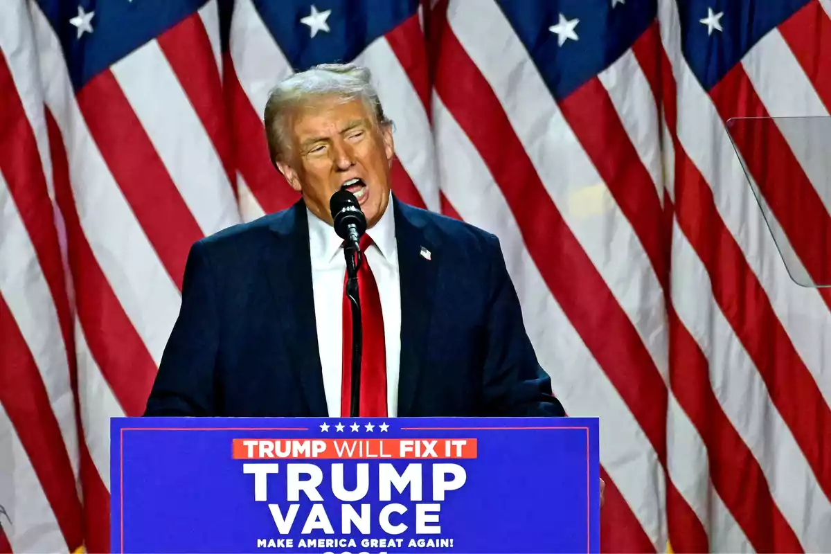 Man in a suit speaking at a podium with United States flags in the background and a sign that says Trump Vance Make America Great Again Man in a suit speaking at a podium with United States flags in the background and a sign that says Trump Vance Make America Great Again