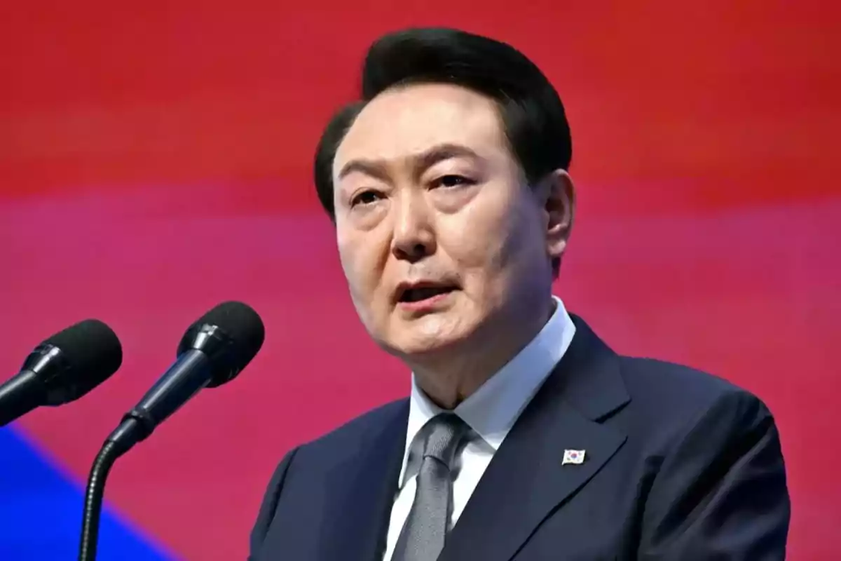 A man in a dark suit and tie speaks in front of two microphones with a red background. A man in a dark suit and tie speaks in front of two microphones with a red background.