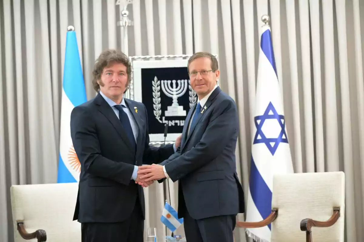 Dos hombres de traje se dan la mano frente a las banderas de Argentina e Israel.