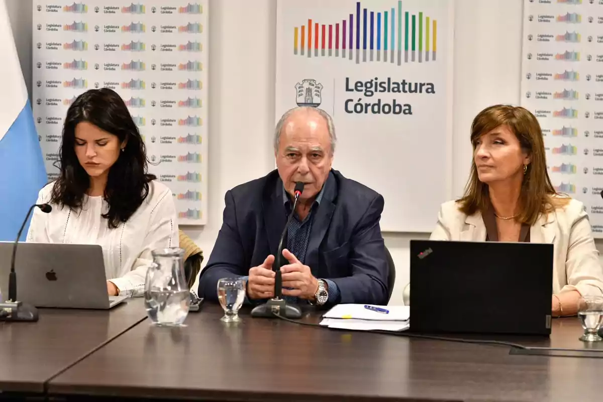 Tres personas sentadas en una mesa con computadoras portátiles y micrófonos, en un entorno de la Legislatura de Córdoba.