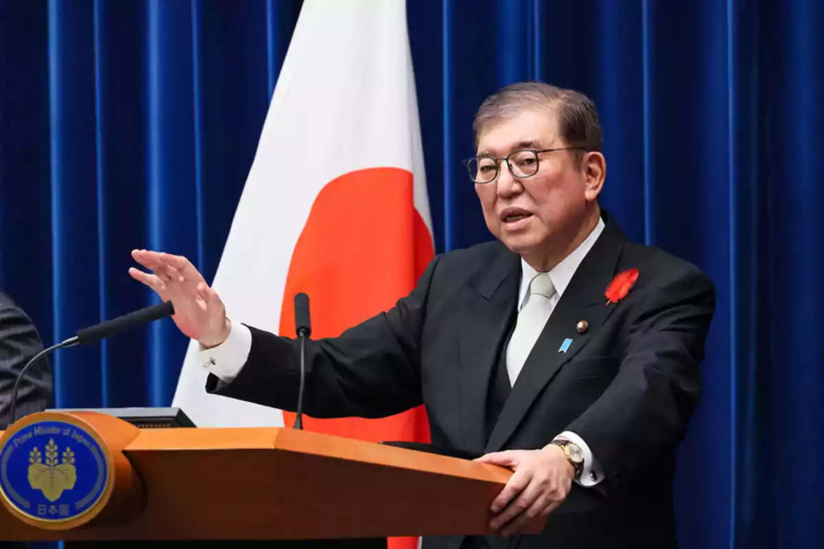 Un hombre en un podio hablando frente a un micrófono con una bandera de Japón de fondo.