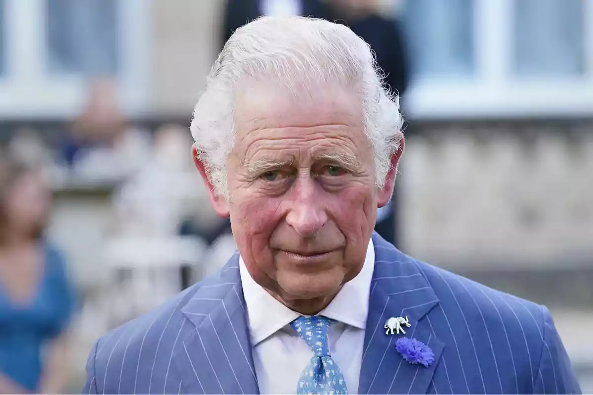 Older man with gray hair and a blue suit with a light blue tie, looking straight ahead in an outdoor setting.
