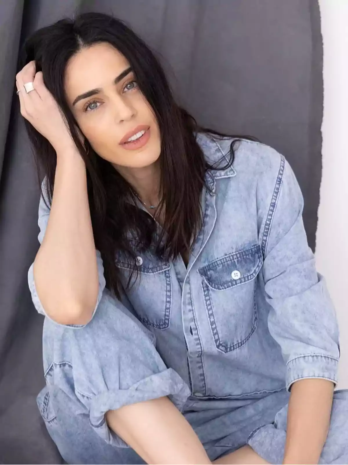 Mujer de cabello oscuro y largo con ropa de mezclilla sentada frente a un fondo gris mirando a la cámara Mujer de cabello oscuro y largo con ropa de mezclilla sentada frente a un fondo gris mirando a la cámara