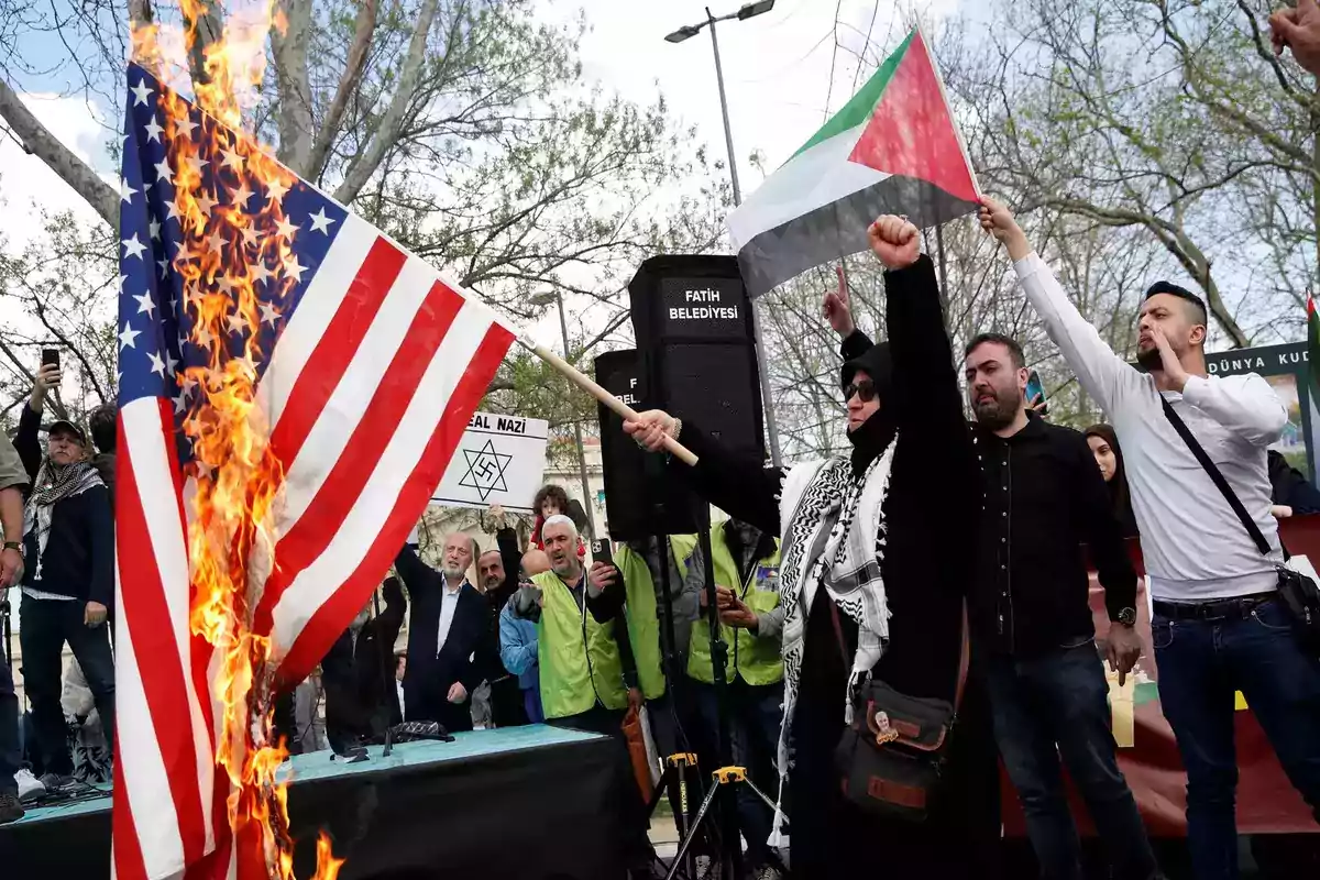 Un grupo de personas participa en una protesta al aire libre donde se quema una bandera de Estados Unidos y se sostiene una bandera palestina.