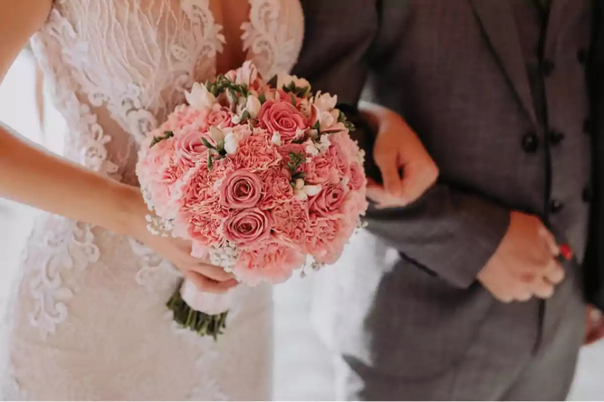 Novia con vestido de encaje sosteniendo un ramo de flores rosas junto a una persona con traje gris.