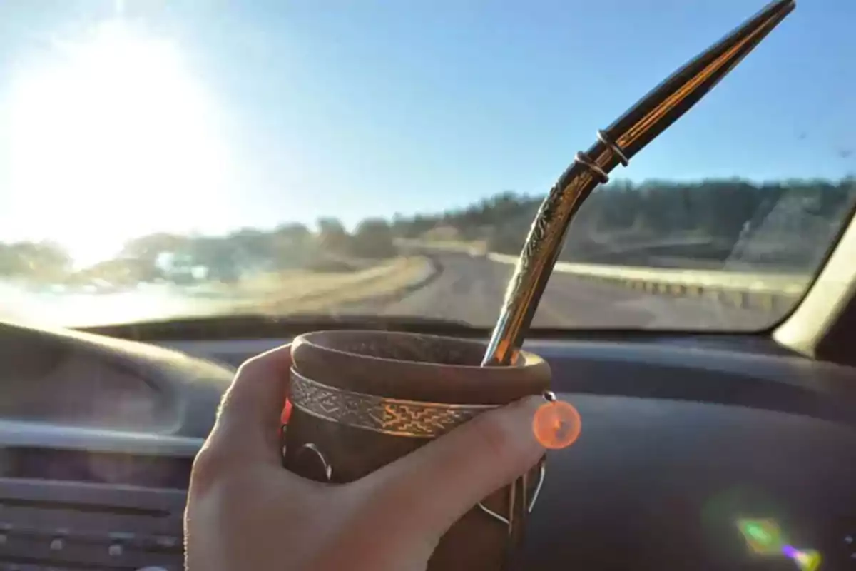 A hand holds a mate with a straw inside a car while observing a road and the sun shining on the horizon. A hand holds a mate with a straw inside a car while observing a road and the sun shining on the horizon.