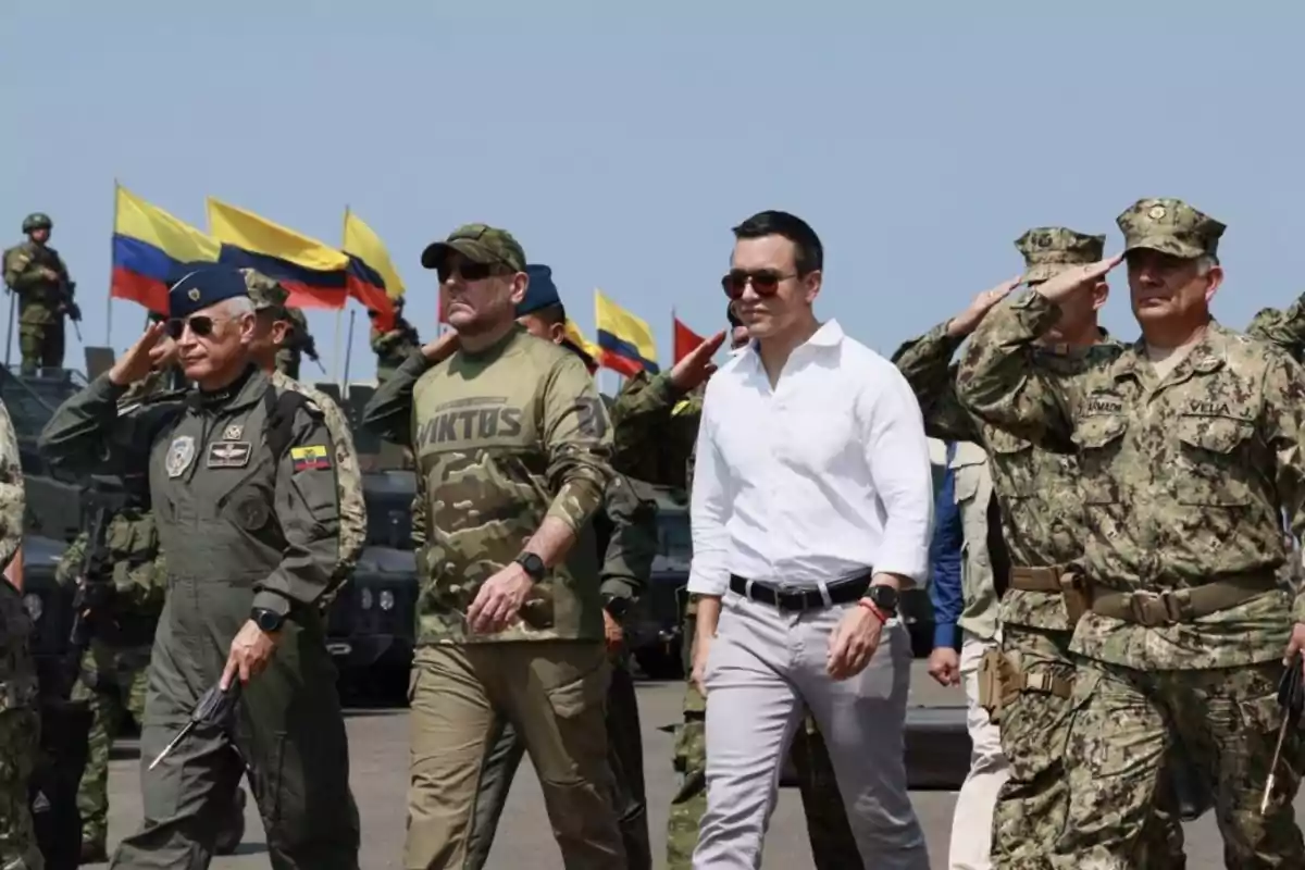 Un grupo de personas en uniforme militar y ropa casual camina mientras algunos saludan, con banderas de Colombia ondeando al fondo.