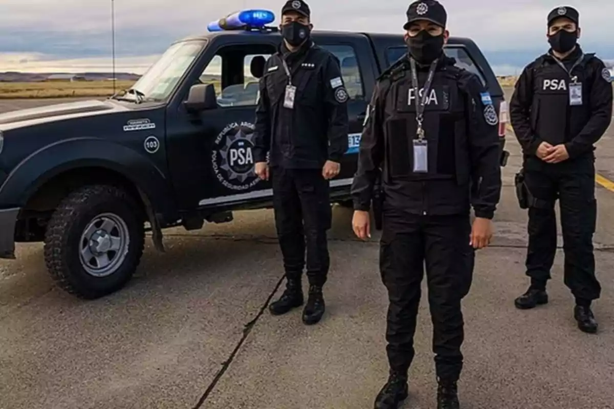 Tres agentes de la Policía de Seguridad Aeroportuaria de Argentina con uniforme negro y mascarillas posan frente a una camioneta oficial en una pista al aire libre