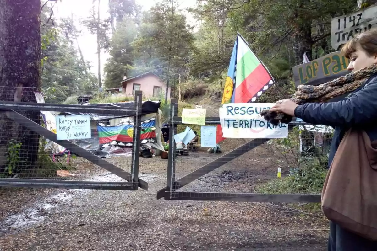 Una persona se encuentra frente a una entrada cercada con varios carteles y una bandera, en un entorno boscoso con una casa al fondo.