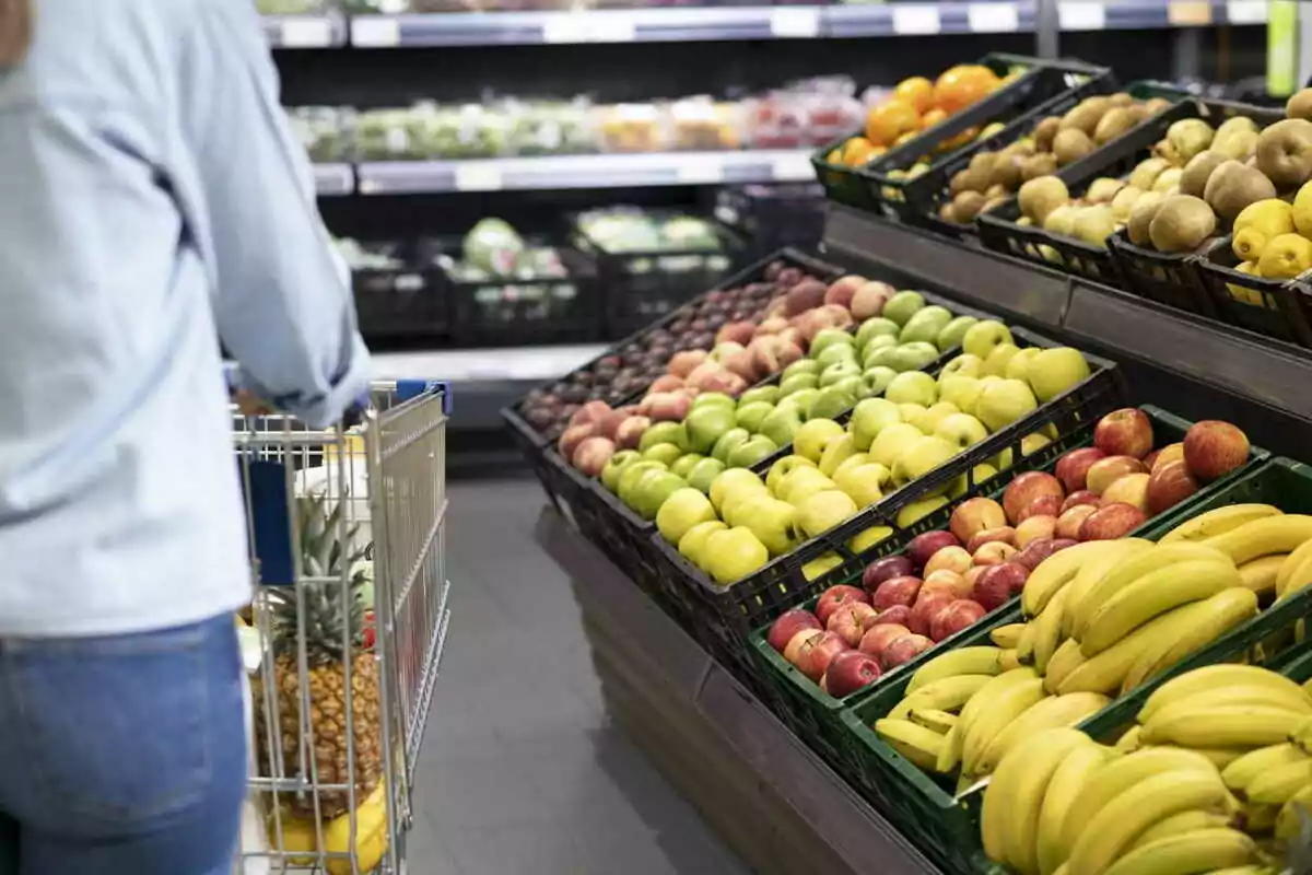 Persona comprando frutas en un supermercado con un carrito lleno de productos.