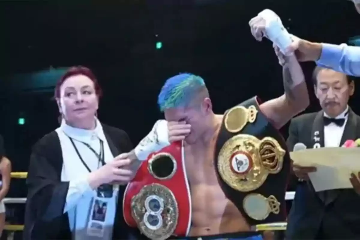 Un boxeador con el cabello teñido de azul y verde está emocionado mientras sostiene varios cinturones de campeonato, acompañado por un árbitro y otra persona en el ring.