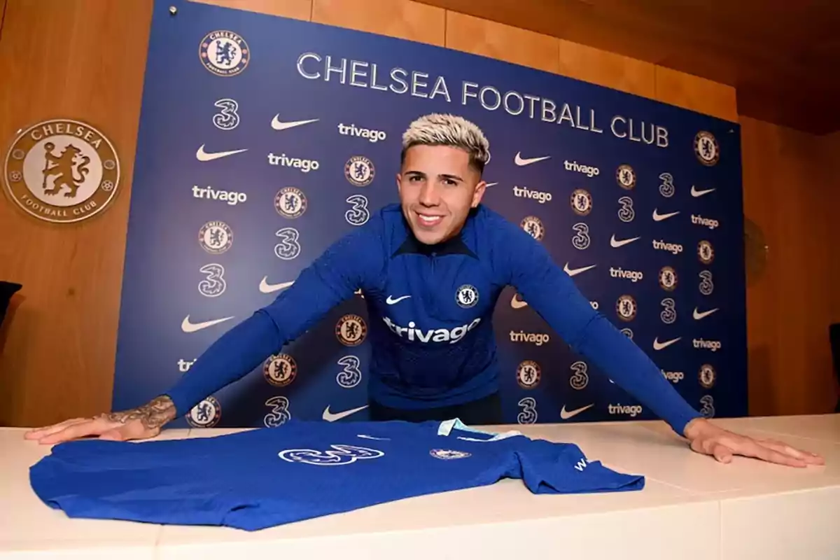 A player in a blue uniform smiles while posing in front of a Chelsea FC jersey on a table, with a background displaying the club's logo and various sponsors. A player in a blue uniform smiles while posing in front of a Chelsea FC jersey on a table, with a background displaying the club's logo and various sponsors.