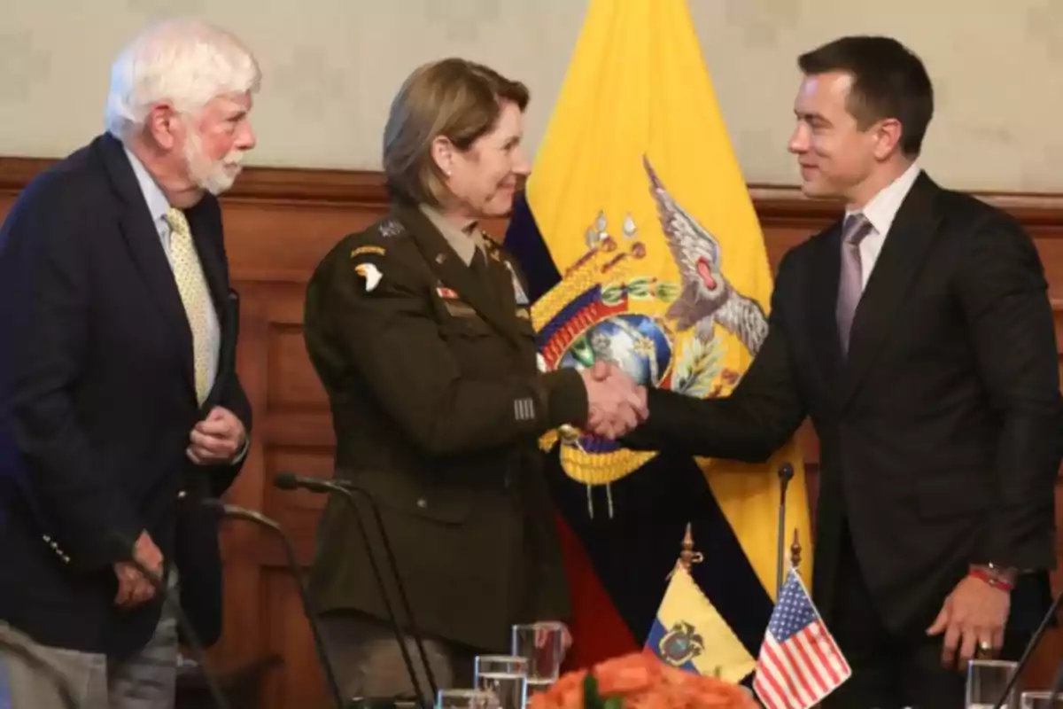 Tres personas en una reunión formal, dos de ellas se dan la mano frente a las banderas de Ecuador y Estados Unidos.