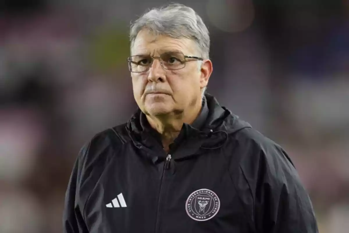 A man with glasses and gray hair is wearing a black jacket with a soccer team logo. A man with glasses and gray hair is wearing a black jacket with a soccer team logo.