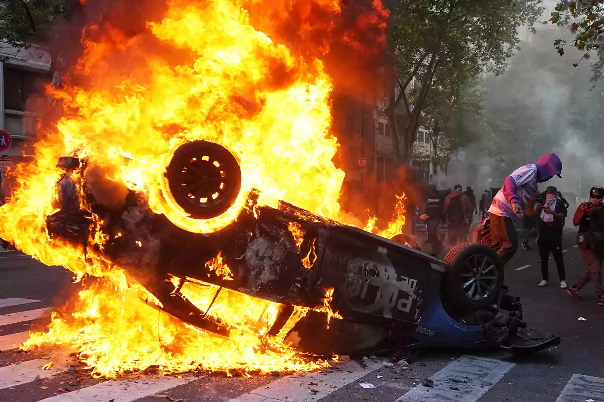 An overturned car in flames on a street with people around.