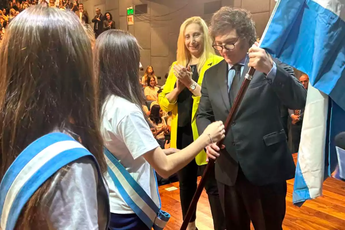 Una ceremonia en la que una persona recibe una bandera de manos de una estudiante, mientras otra persona aplaude en el fondo.