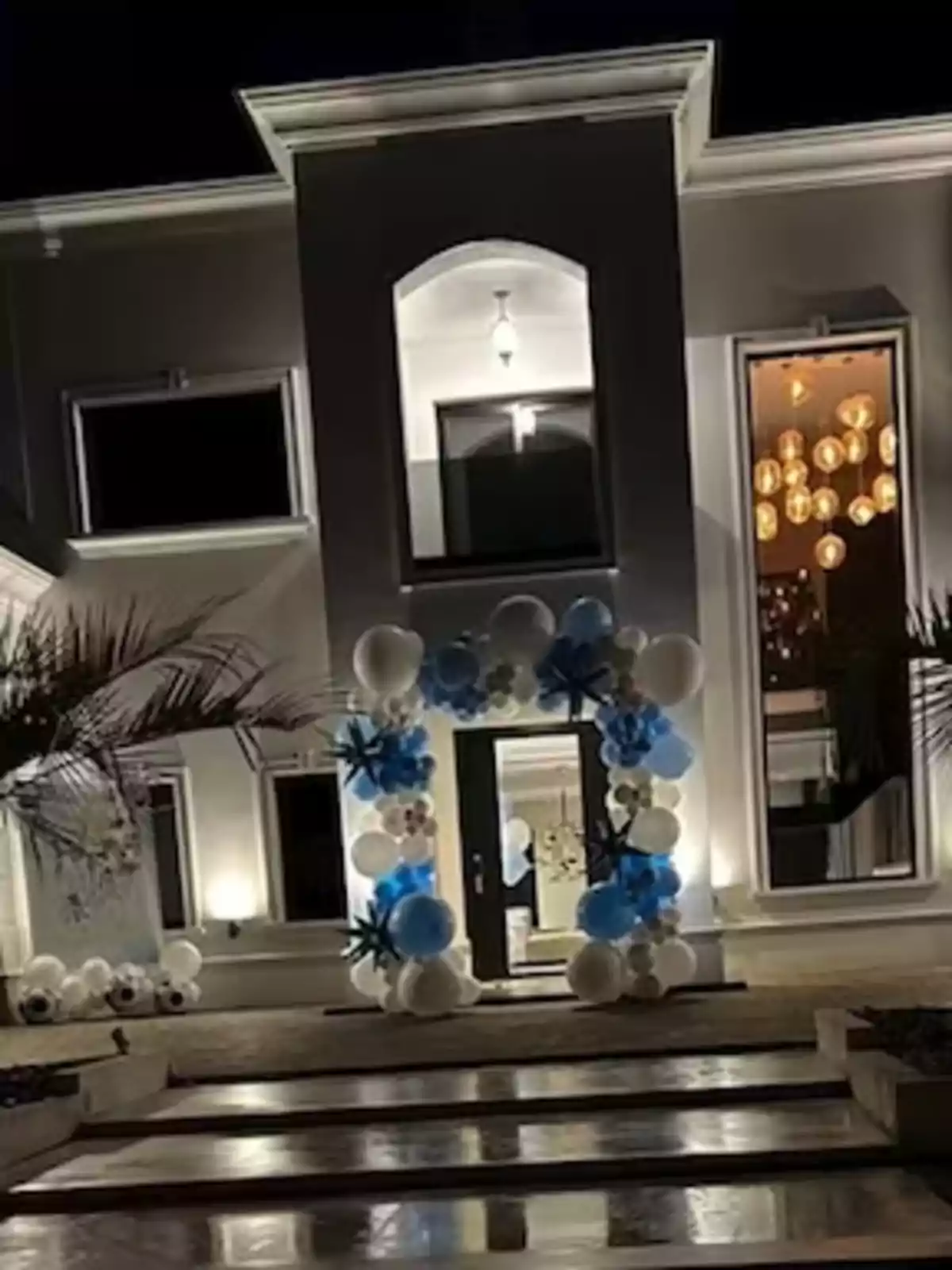 Una casa moderna iluminada por la noche con una entrada decorada con un arco de globos blancos y azules.