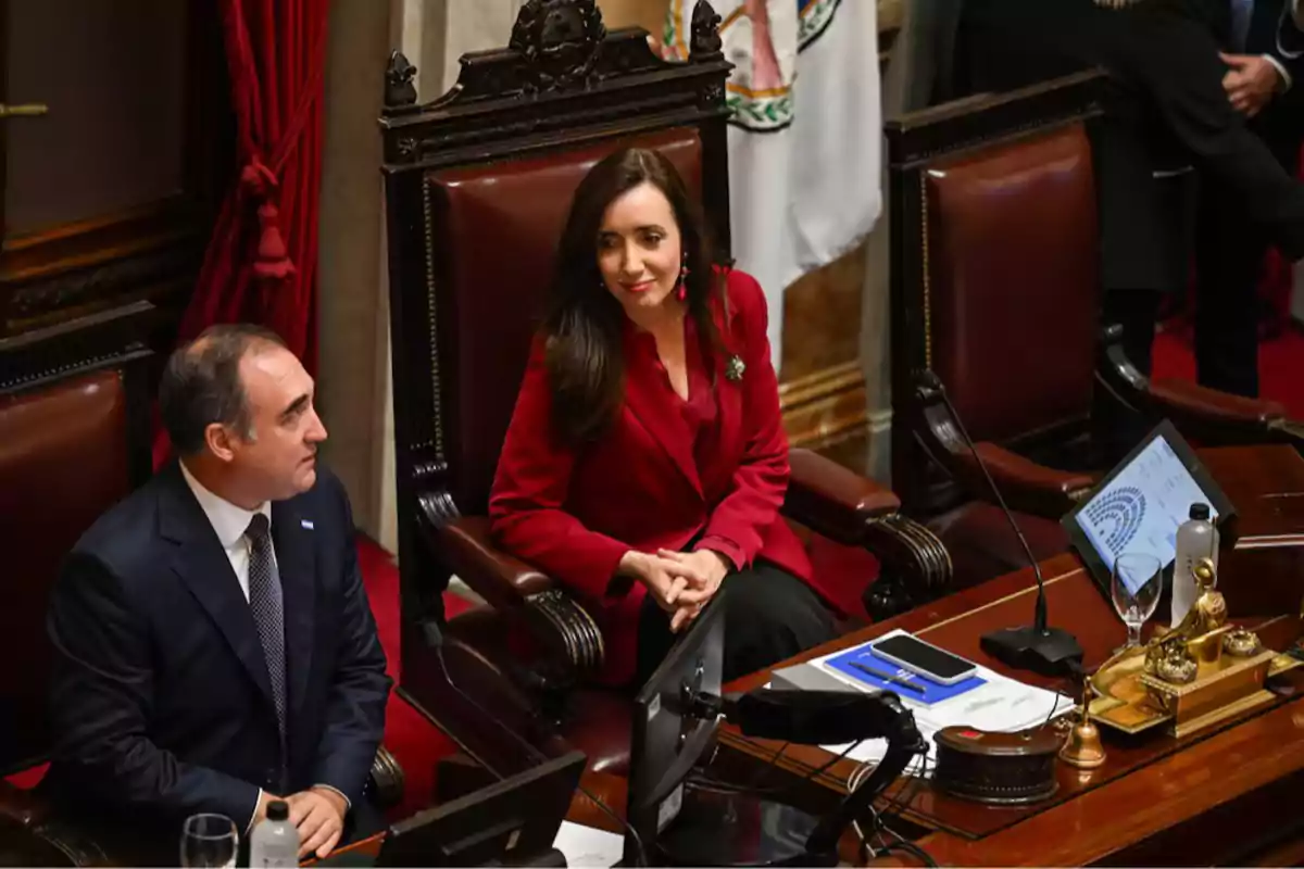 Dos personas sentadas en sillas de madera en un entorno formal con documentos y dispositivos electrónicos sobre la mesa. Dos personas sentadas en sillas de madera en un entorno formal con documentos y dispositivos electrónicos sobre la mesa.