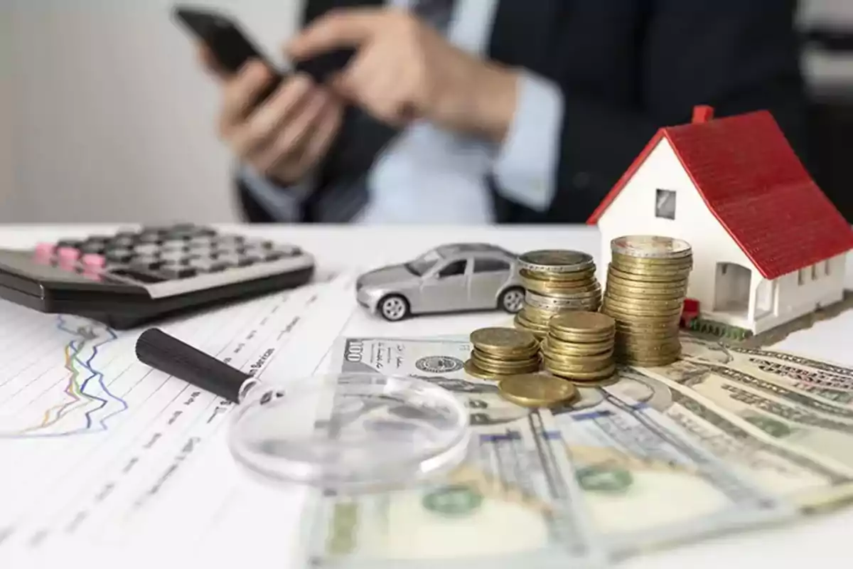 Monedas apiladas, billetes, una casa y un auto en miniatura sobre una mesa con gráficos, una lupa, una calculadora y una persona usando un teléfono móvil al fondo