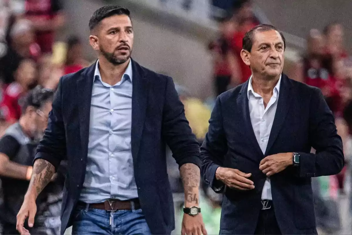 Two men in dark jackets and light jerseys walk together in a soccer stadium with blurred people in the background. Two men in dark jackets and light jerseys walk together in a soccer stadium with blurred people in the background.