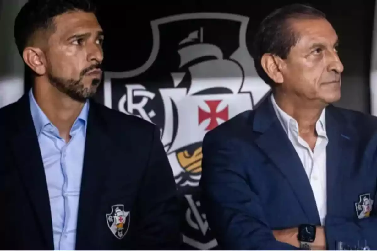 Two men wearing blue blazers and light-colored jerseys look in opposite directions with the Vasco da Gama club crest in the background.