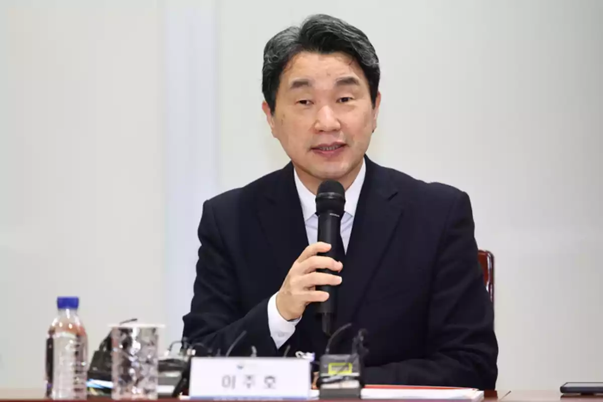 A man in a dark suit speaks into a microphone during a press conference, with water bottles and papers on the table. A man in a dark suit speaks into a microphone during a press conference, with water bottles and papers on the table.