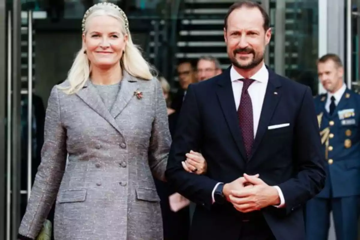 A smartly dressed couple walks hand in hand in front of a building with people in the background.